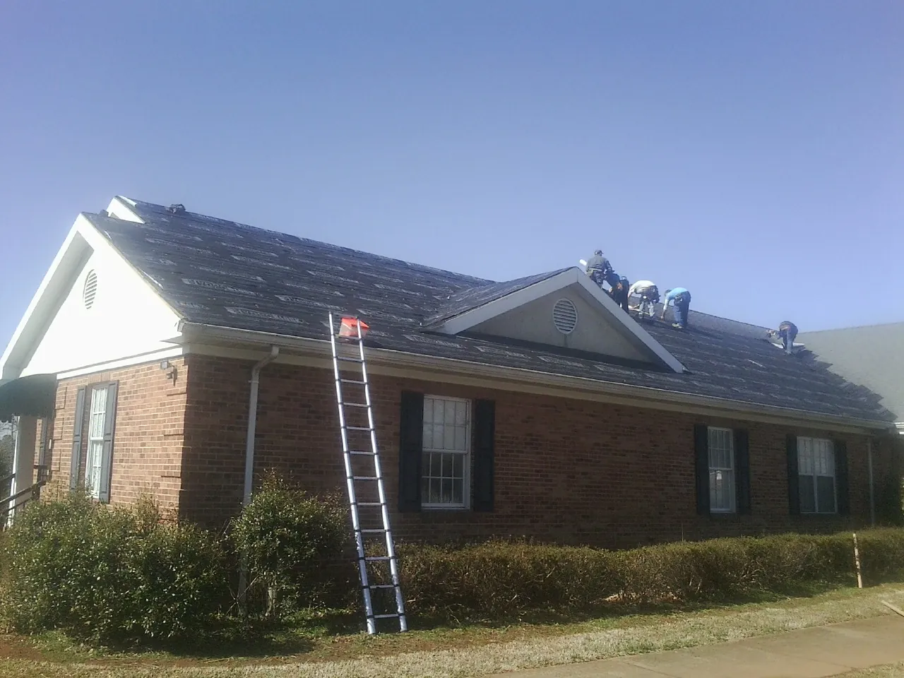 Crew tearing shingles off a brick ranch roof with a ladder set — Emergency Roof Repair work by our crew in Rogerson