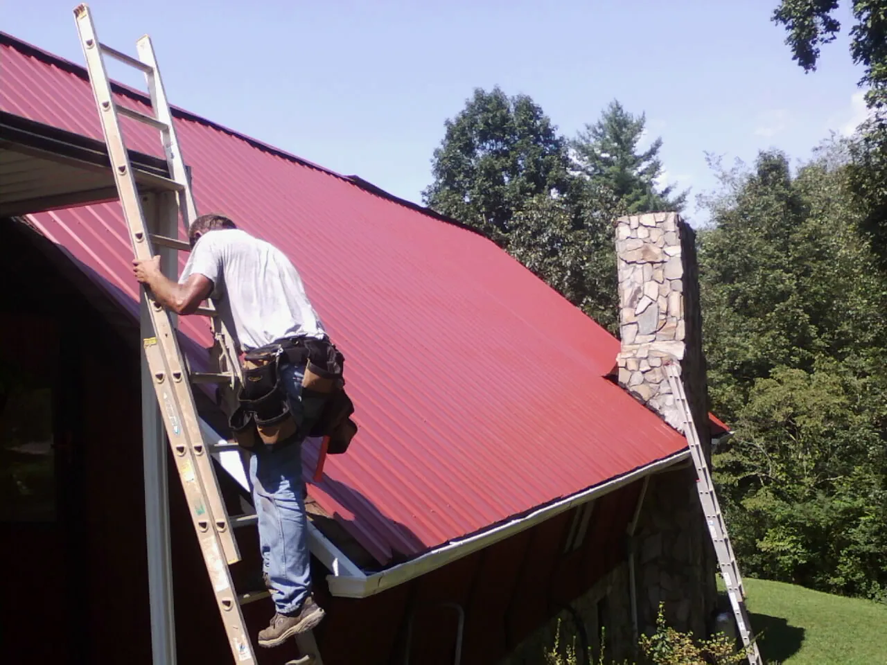 Roofer on a ladder beside a red metal roof and stone chimney — Soffit & Fascia Repair by Roofing Brothers in Rogerson